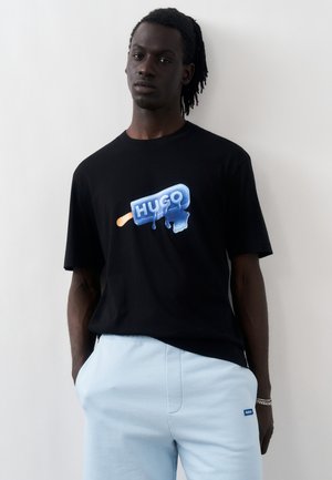 Μαύρο μπλουζάκι με μπλε λιωμένο παγωτό γραφικό με την επιγραφή "HUGO", που φοριέται από άνδρα με τα χέρια στις τσέπες, συνδυασμένο με ανοιχτό μπλε παντελόνι.