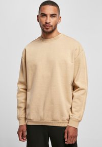 Sudadera beige con cuello redondo, corte holgado y puños acanalados. Textura de tela suave sin patrones visibles ni detalles de herrajes.