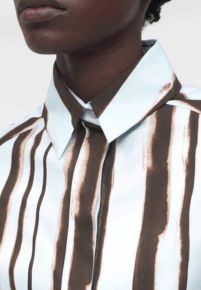 Κοντινό πλάνο ενός ατόμου που φοράει λευκό πουκάμισο με ακανόνιστες κάθετες καφέ ρίγες και μυτερό γιακά, εστιάζοντας στον λαιμό και το πάνω μέρος του στήθους.