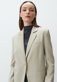 Beige Blazer mit einem einzigen Knopf, Revers und schlanker Silhouette. Verfügt über eine Brusttasche und eine glatte Stoffstruktur.