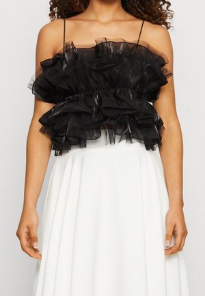 Vrouw draagt een zwarte, geruwde, strapless crop top met dunne bandjes en een witte geplooide rok met hoge taille tegen een effen achtergrond.