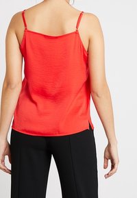 Top de satén rojo con tirantes finos y ajustables, que presenta un dobladillo recto y aberturas laterales para facilitar el movimiento. Textura suave y ligero.