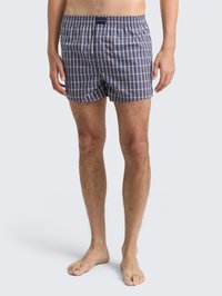 Man in blauw en grijs geruite boxershorts die blootsvoets op een lichtgrijze achtergrond staat.