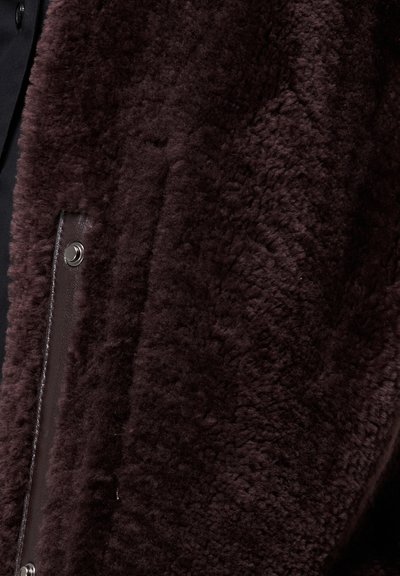 Brūna mākslīgās kažokādas jaka ar mīkstu tekstūru, ar aizdari ar spraudņiem un ādas apdari. Dizainā ir brīvs piegriezums un minimāla dekorācija.