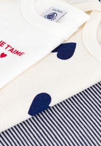 Drei Stück Kinderbekleidung: eines weiß mit rotem Text und Herz, eines cremefarben mitnavyblauen Herzen und eines mit navyblauen Streifen.