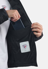 Giacca puffer nera con una tasca con zip. L'etichetta bianca all'interno presenta un logo. Una mano tiene uno smartphone, mostrando il design della tasca.