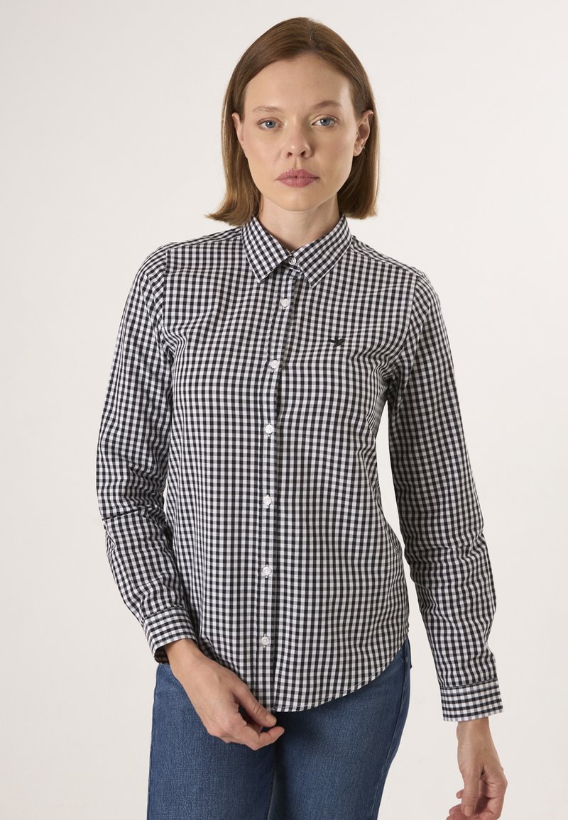 Femme portant une chemise à carreaux noire et blanche à manches longues et un jean bleu, debout devant un fond clair uni.