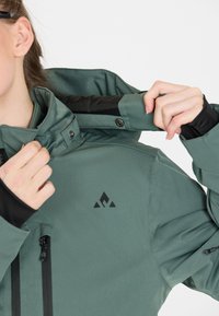 Grüne wasserdichte Jacke mit schwarzen Akzenten, hohem Kragen, verstellbaren Funktionen und einem Logo auf der Brust. Sichtbare Reißverschlusstaschen.