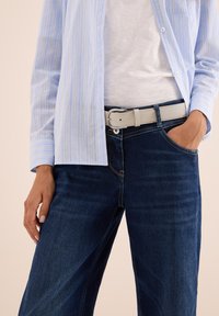Ljusblå och vit randig skjorta med knappar över en vit t-shirt, kombinerat med mörkblå jeans och ett ljusgrått bälte, hand i fickan.