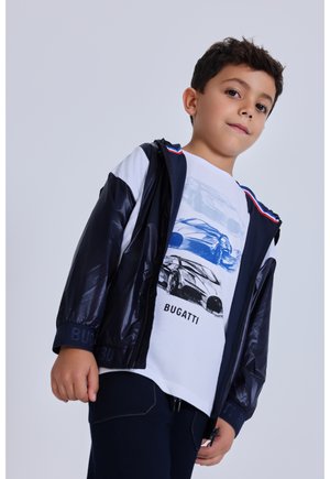 Junge trägt eine marineblaue Jacke über einem weißen Bugatti-T-Shirt mit Autoskizzen, steht vor einem schlichten hellen Hintergrund und blickt leicht nach unten.
