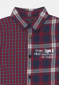 Blau-rot kariertes Hemd mit Button-Down-Kragen, weißen Karomustern und gesticktem Text, aus Baumwollstoff gefertigt.