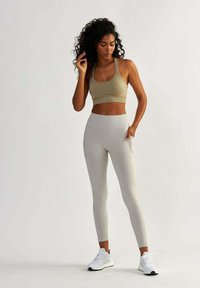 Reggiseno sportivo beige e leggings grigi chiari con tasche laterali. Modella che indossa scarpe da ginnastica bianche, in piedi su uno sfondo neutro.