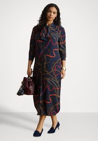 Sieviete ar cirtainiem matiem, valkājot tumši zilu midi kleitu ar rakstu, turot bordozas krāsas rokassomiņu, tumši zilos papēžus, zelta aproci un riņķa formas auskarus.