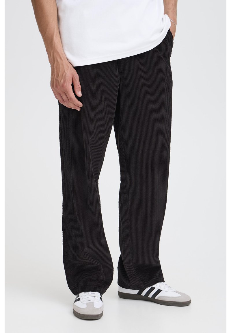 Pantalones de pana negros con un corte ancho y relajado y una textura acanalada vertical; combinados con camiseta blanca y zapatillas grises con rayas negras.