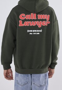 Vihreä huppari, jossa suuri punainen teksti "Soita asianajajalleni" selässä, valkoisella puhelinnumerolla ja aukioloajoilla. Pehmeä materiaali, löysä istuvuus.