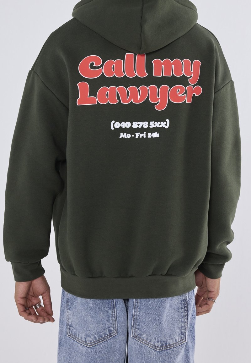 Vihreä huppari, jossa suuri punainen teksti "Soita asianajajalleni" selässä, valkoisella puhelinnumerolla ja aukioloajoilla. Pehmeä materiaali, löysä istuvuus.