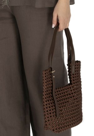 Ręka trzyma ciemnobrązową plecioną torebkę z skórzanymi paskami, na tle brązowych spodni.