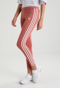 Ljusrosa leggings med hög midja, med tre vita sidostrekar och en adidas-logotyp. Tillverkade av elastisk kvalitet med jämn textur.