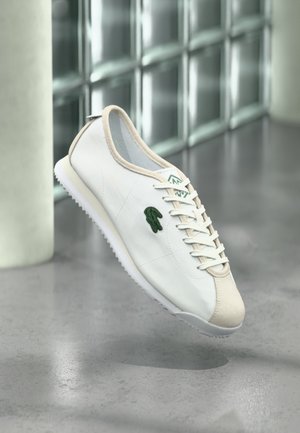 Zapatilla blanca con ribete beige y logotipo de cocodrilo verde, mostrada flotando sobre un suelo gris cerca de paneles de pared de vidrio esmerilado.