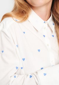 Camicia bianca con motivi a cuore blu, colletto classico e chiusura a bottoni. Tessuto testurizzato con accenti ricamati in tutta la superficie.