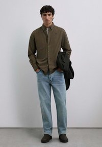 Camisa de botones con un patrón de cuadros en verde oscuro, combinada con unos vaqueros de color azul claro. Un blazer negro colgado del brazo y calzado oscuro completan el conjunto.