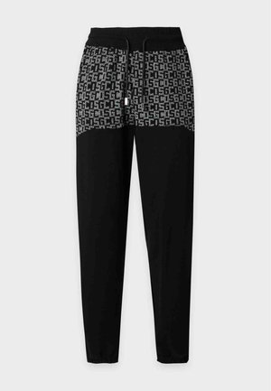 Jeune homme portant une chemise à manches courtes noire et grise à motifs et un pantalon assorti avec des baskets blanches, debout devant un fond blanc.