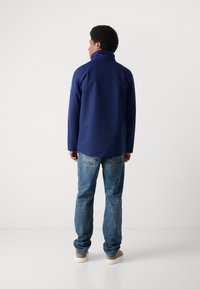 Veste imperméable bleu marine avec un col haut, présentant une texture lisse et un design minimaliste, associée à un jean en denim bleu clair et des chaussures beiges.
