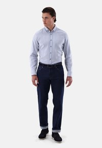 Gestreifte Hemdjacke in Hellblau und Weiß, kombiniert mit dunkelblauen Jeans. Das Modell steht mit den Händen an den Seiten und trägt schwarze Schuhe.