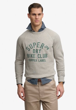 Man draagt een lichtgrijze trui met de tekst 'Super Dry Bike Club Copper Label', een blauw geruit overhemd, beige broek en een horloge aan de linkerpols.