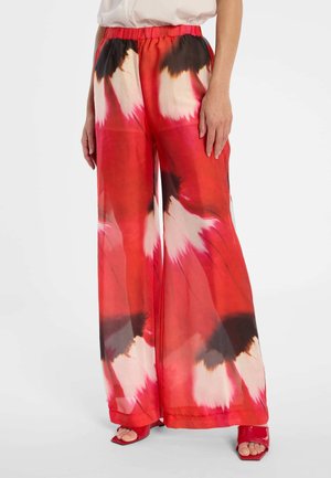 Weit geschnittene Hose mit abstraktem Blumenmuster in Rot, Weiß und Schwarz, getragen mit roten offenen Schuhen auf weißem Hintergrund.