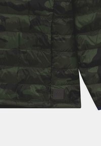 Giacca puffer verde mimetica con texture trapuntata, dotata di una patch logo GAP grigio scuro in basso a destra. Presenta una finitura lucida.