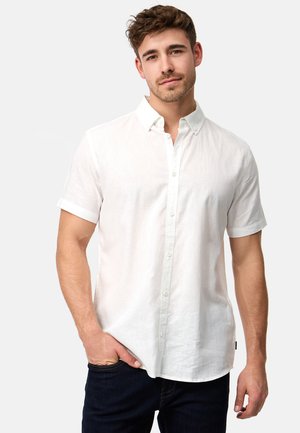Giovane uomo con capelli corti castani che indossa una camicia bianca a maniche corte con bottoni e jeans scuri, in piedi con una mano in tasca.