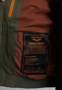 Interior de chaqueta de vuelo de cuero verde oliva con forro marrón. Presenta detalles en naranja en el cierre, etiqueta y bolsillos; puños y cinturilla acanalados.