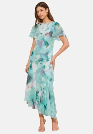 Kvinde iført en flydende kjole til anklerne med grøn-blå akvarelblomster og flagrende ærmer, kombineret med beige sandaler med høj hæl.