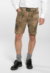 Lederhosen en cuir couleur tan, ornés de broderies décoratives, d'une ceinture avec une boucle en métal et de boutons sur les côtés. Associés à des chaussettes grises enroulées et des chaussures noires.