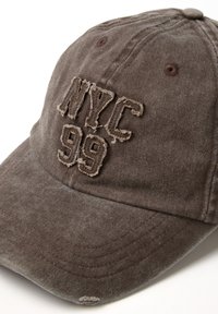 Cappellino da baseball in cotone marrone usurato con patch ricamato sfilacciato che legge "NYC 99" sul pannello anteriore, su sfondo bianco.