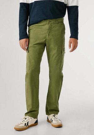 Olivgrüne Cargo-Hosen mit mehreren Seitentaschen, Gürtelschlaufen, geradem Schnitt und aus Baumwollstoff. Kombiniert mit weißen und marineblauen Turnschuhen.
