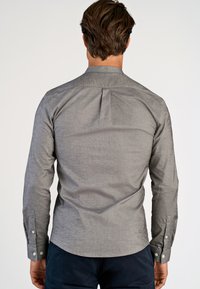 Uomo che indossa una camicia a maniche lunghe grigio chiaro con bottoni e pantaloni scuri, visto da dietro su uno sfondo semplice.