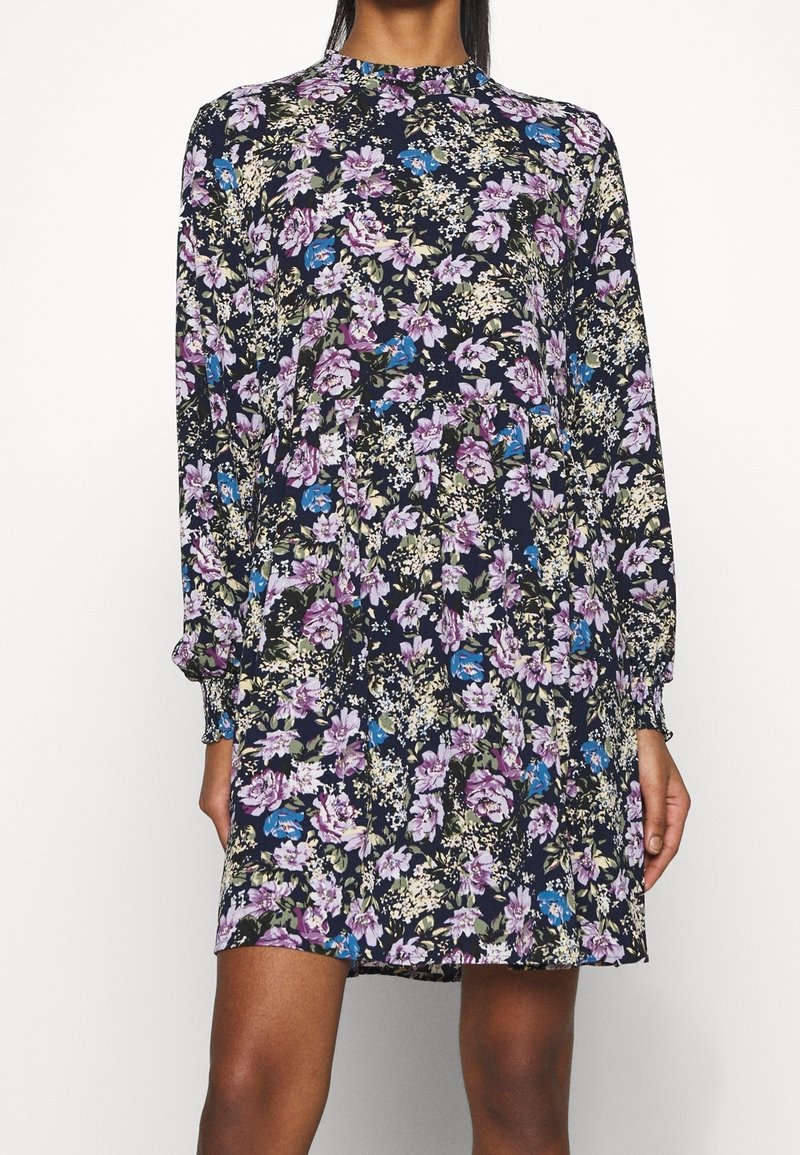 Femme portant une robe à manches longues, longueur genou, avec un col montant et un motif floral foncé avec des fleurs violettes, bleues et blanches.