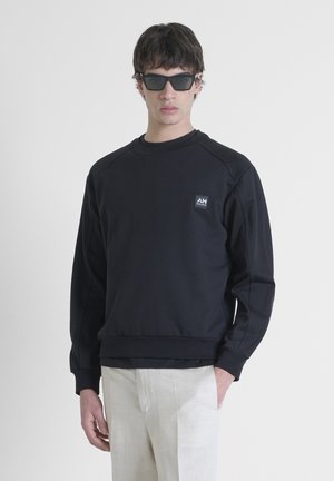 Sudadera negra con cuello redondo, mangas raglán y un pequeño parche con el logo en el lado izquierdo del pecho. Textura suave y corte relajado.
