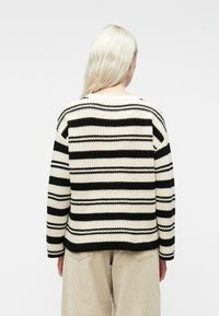 Pikkade blondide juustega inimene, seljaga kaamera poole, musta ja kreemika horisontaalselt triibulise kudumiga ning beežide pükstega, lihtsal taustal.