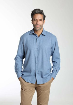 Homme aux cheveux gris et barbe, portant une chemise bleu clair à boutons et un pantalon beige, debout les mains dans les poches devant un fond blanc.
