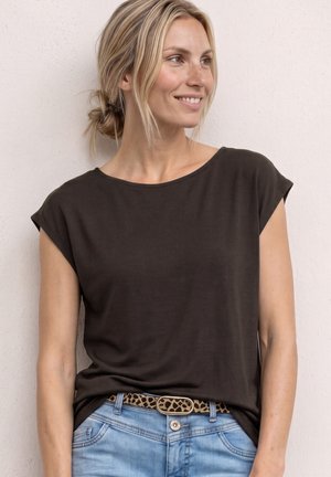 Femme aux cheveux blonds en chignon bas, portant un haut marron foncé à manches courtes, un jean bleu clair et une ceinture imprimé léopard, regardant vers la droite.