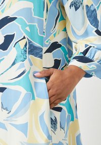 Langärmliges Kleid aus leichtem Stoff mit einem blauen, weißen und gelben Blumenmuster, seitlicher Tasche und gerafftem Ärmelabschluss.