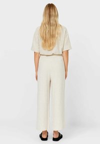Donna con lunghi capelli biondi ondulati, che indossa una maglietta beige a maniche corte e pantaloni a gamba larga, in piedi a piedi nudi su uno sfondo bianco.
