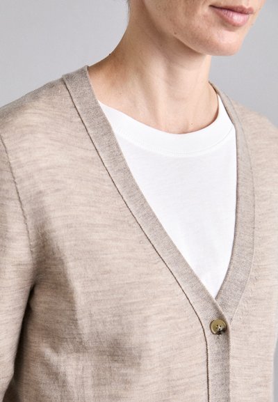 Beige Cardigan mit V-Ausschnitt, aus weichem Stoff. Verfügt über einen einzigen Knopfverschluss und ein weißes T-Shirt darunter. Glatte Textur.