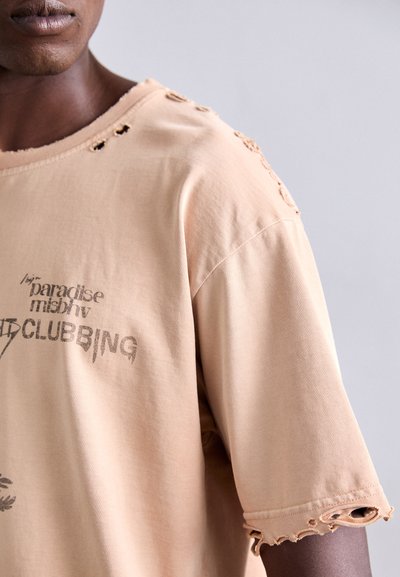 Nærbillede af en person, der bærer en beige slidt t-shirt med trykt tekst, som viser slidte huller på skulderen og ærmespidserne.