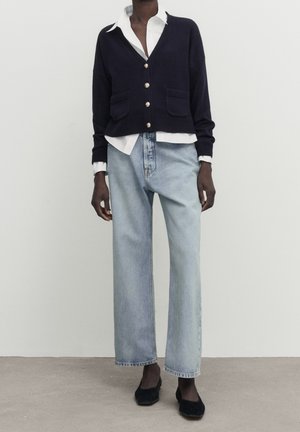 Osoba ubrana w granatowy kardigan na guziki, białą koszulę z kołnierzykiem, jasnoniebieskie szerokie dżinsy i czarne baleriny, stojąca na tle gładkiej ściany.