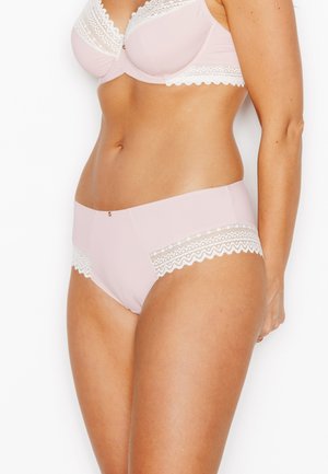 Femme portant un soutien-gorge rose pâle et une culotte assortie avec une bordure en dentelle blanche, debout sur un fond blanc.