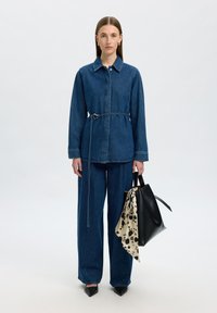 Giacca di jeans con maniche lunghe e cintura in vita, abbinata a pantaloni di jeans larghi. Tiene in mano una borsa nera con una sciarpa crema con macchie nere.